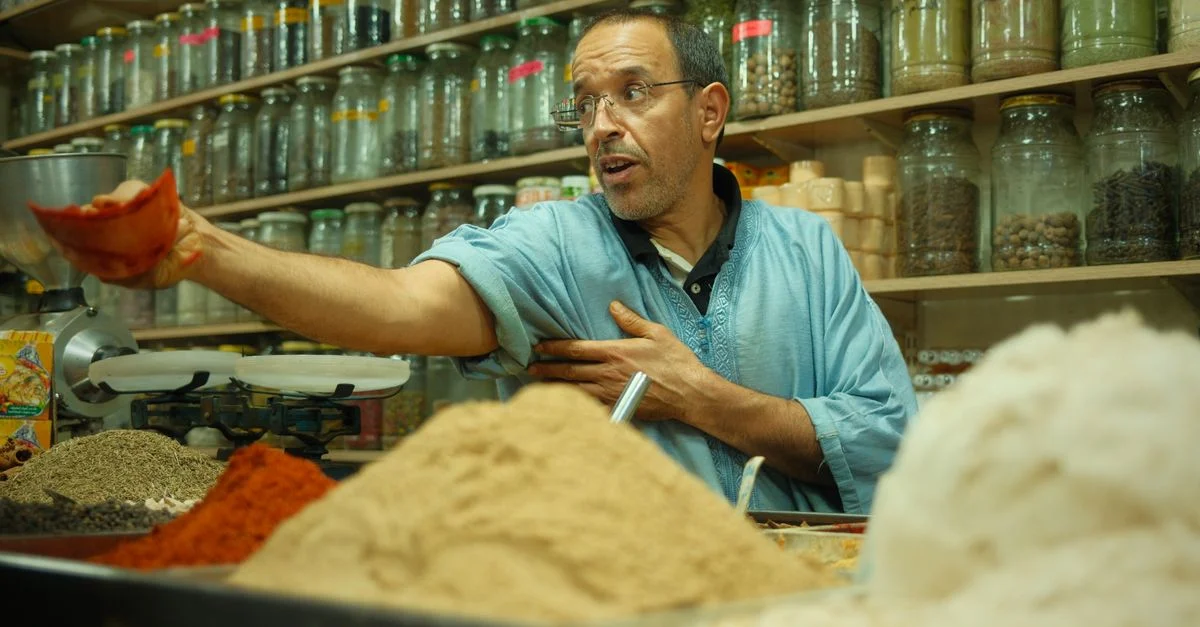 Un homme marocain vendant des épices dans une étal coloré de marché traditionnel, présentant diverses herbes et épices.