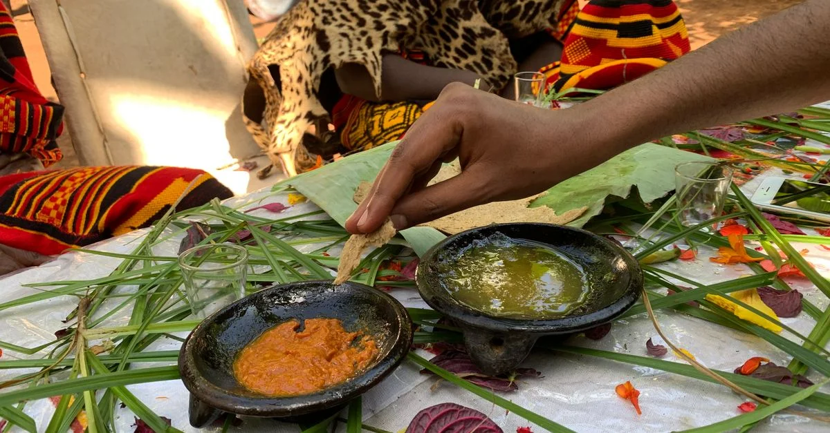 Préparation à la main d'un plat africain avec deux sauces au milieu de textiles traditionnels colorés.