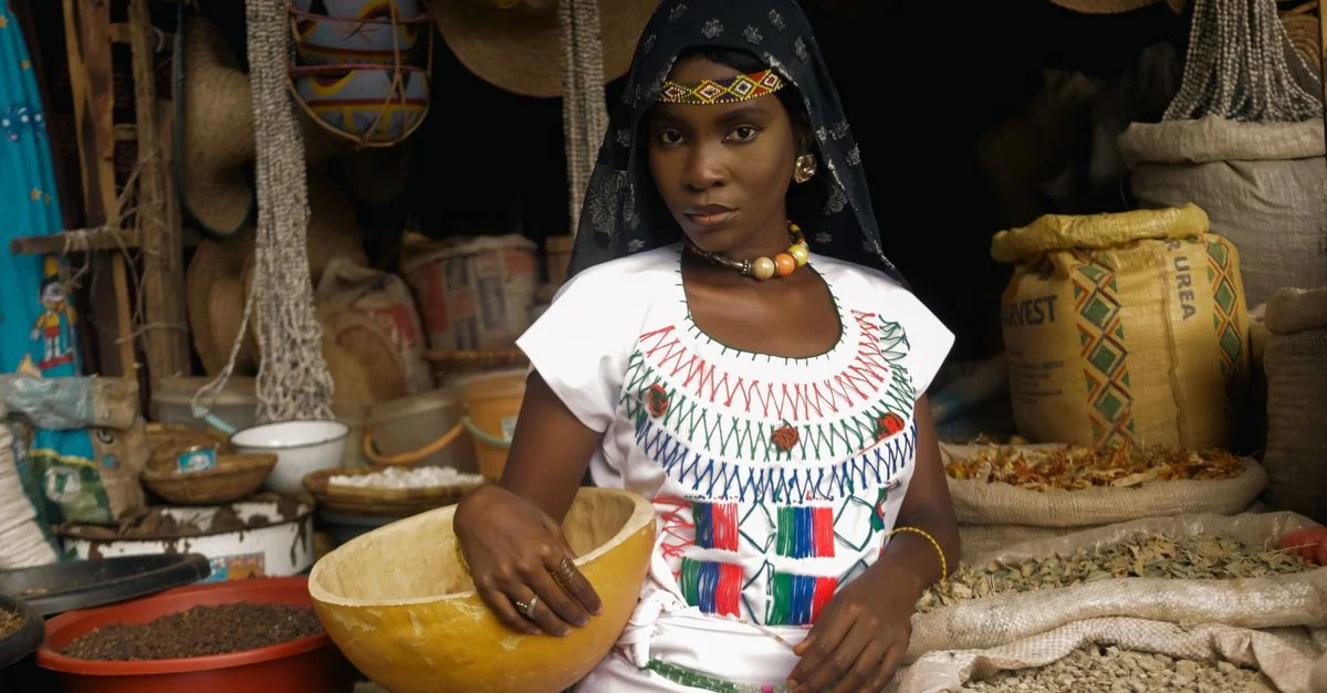 Une vendeuse en tenue traditionnelle à un étal de marché africain rempli de textiles colorés et de grains.