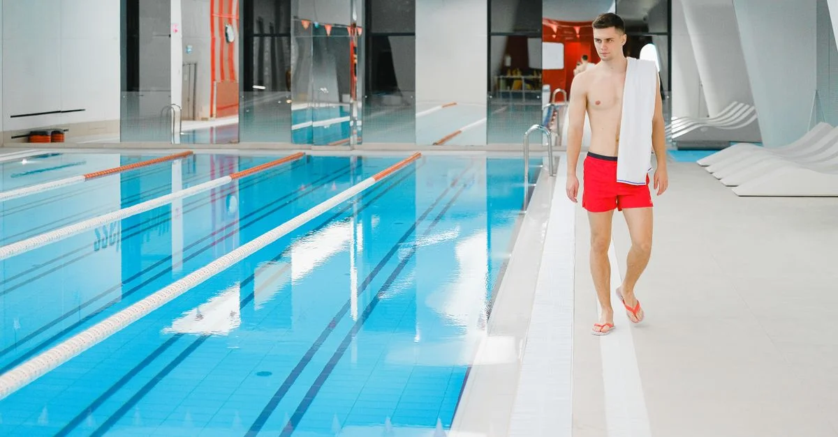 Homme jeune torse nu marchant au bord de la piscine dans un complexe aquatique intérieur avec une serviette sur l'épaule.